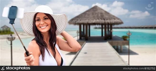lifestyle, leisure, summer, technology and people concept - smiling young woman or teenage girl in sun hat taking picture with smartphone on selfie stick over bungalow on beach background