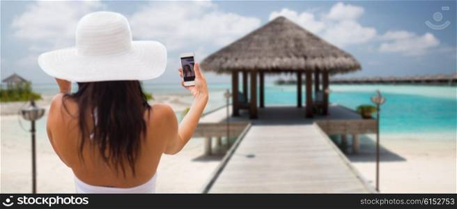 lifestyle, leisure, summer, technology and people concept - smiling young woman or teenage girl in sun hat taking selfie with smartphone over bungalow on beach background