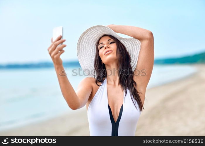 lifestyle, leisure, summer, technology and people concept - smiling young woman or teenage girl in sun hat taking selfie with smartphone on beach