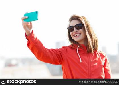 lifestyle, leisure, technology and people concept - smiling young woman or teenage girl in sunglasses taking selfie with smartphone outdoors