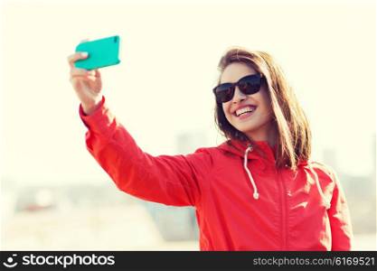 lifestyle, leisure, technology and people concept - smiling young woman or teenage girl in sunglasses taking selfie with smartphone outdoors