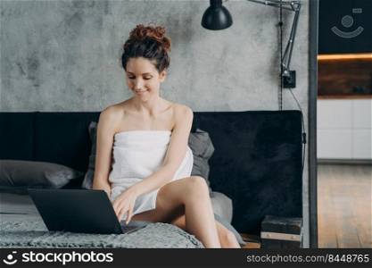 Lifestyle of a freelancer. Attractive spanish girl wrapped in towel after bathing is sitting at laptop on her bed and smiling. Young woman takes shower and relaxing. Remote worker at home.. Freelancer spanish girl wrapped in towel after bathing is sitting at laptop on her bed and smiling.