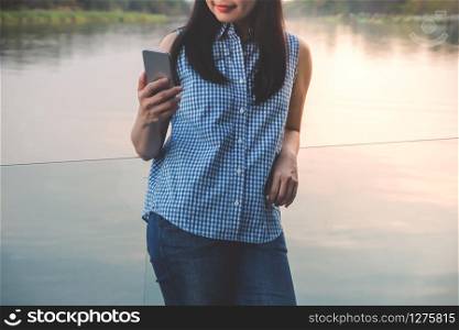 Lifestyle of Modern People Concept. Young Woman Relaxing by Reading Data or Message via Smartphone. Standing at the Deck by Riverside in Summer