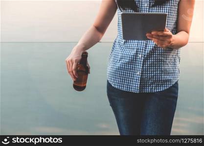 Lifestyle of Modern People Concept. Young Woman Relaxing by Reading Data via Tablet and Drinking Beer. Standing at the Deck by Riverside in Summer