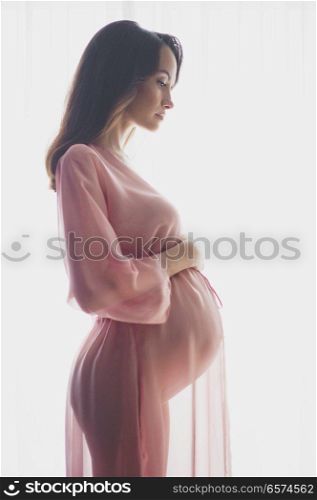 Lifestyle portrait of beautiful pregnant woman in pink negligee. Beautiful morning. Beauty and health