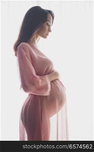 Lifestyle portrait of beautiful pregnant woman in pink negligee. Beautiful morning. Beauty and health