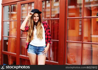 Lifestyle portrait of fashionable blonde girl with ombre hair wearing a rock red shirt, black cap, white t-shirt and denim shorts having fun outdoors in the city. Swag style.