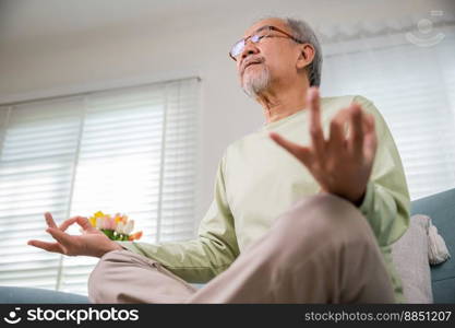 Lifestyle senior man sitting on sofa in living room holding hands in mudra practicing home yoga in lotus pose, Elderly relaxed man do physical exercises following healthy on stress free weekend