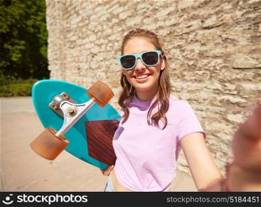 lifestyle, summer, longboarding and people concept - smiling young woman or teenage girl in sunglasses with longboard taking selfie over stone wall outdoors. teenage girl with longboard taking selfie outdoors