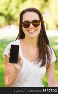 lifestyle, summer vacation, advertisement, technology and people concept - smiling young girl showing black blank smartphone screen and sitting on grass in park