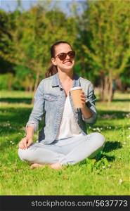 lifestyle, summer vacation, drinks and people concept - smiling young girl drinking coffee from paper cup and sitting on grass in park