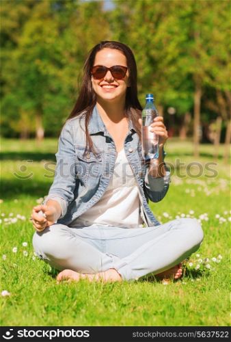 lifestyle, summer, vacation, drinks and people concept - smiling young girl in sunglasses with bottle of water sitting on grass in park