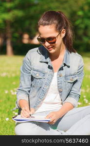 lifestyle, summer vacation, education and people concept - smiling young girl writing with pencil to notebook and sitting on grass in park