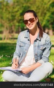 lifestyle, summer vacation, education and people concept - smiling young girl writing with pencil to notebook and sitting on grass in park