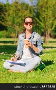 lifestyle, summer vacation, education, drinks and people concept - smiling young girl writing to notebook and drinking coffee in park
