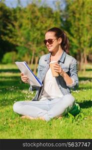 lifestyle, summer vacation, education, drinks and people concept - smiling young girl reading notes from notebook and drinking coffee in park