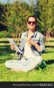 lifestyle, summer vacation, education, drinks and people concept - smiling young girl reading notes from notebook and drinking coffee in park