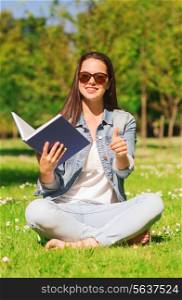 lifestyle, summer vacation, education, gesture and people concept - smiling young girl with book showing thumbs up and sitting in park