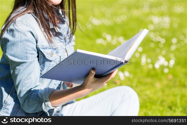 lifestyle, summer vacation, education, literature and people concept - close up of smiling young girl reading book and sitting on grass in park