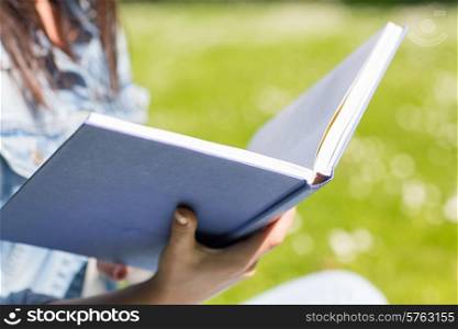 lifestyle, summer vacation, education, literature and people concept - close up of young girl reading book and sitting on grass in park