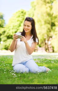 lifestyle, summer, vacation, technology and people concept - smiling young girl with smartphone sitting on grass in park