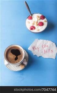 Light breakfast with fresh Arabic coffee and a bowl of yogurt with cereal and fruits and a piece of paper with the pay the bills message, on a blue table, in the morning light.