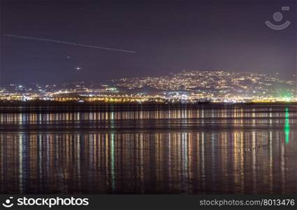 Light streak in sky over city at night. Long light streak from airplane or meteor in sky over city on hill with coastline in foreground at night