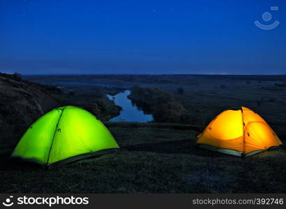 Lighted from the inside orange and green tents on a hill above the river. Night landscape. The concept of freedom, privacy and travel.. Lighted from the inside orange and green tents on hill above riv
