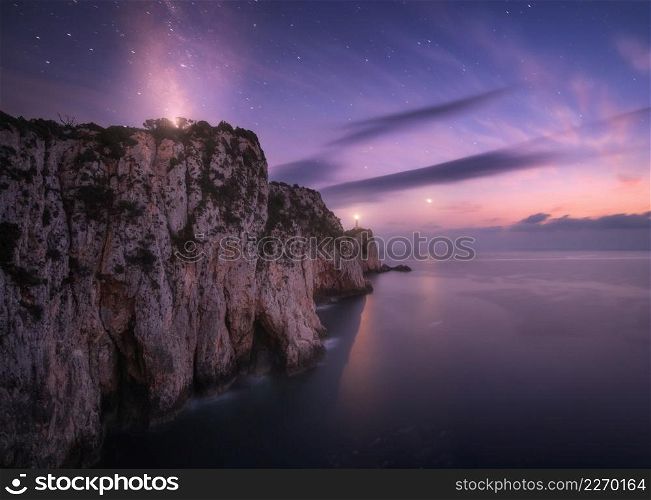 Lighthouse on the mountain peak at starry night in summer. Beautiful cliffs, rocky sea coast, bright stars, milky way and violet sky with clouds at dusk. Lighthouse of Cape Lefkada, Greece. Landscape