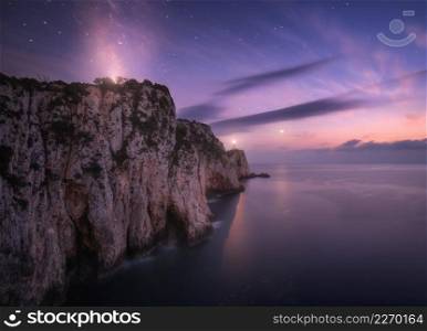 Lighthouse on the mountain peak at starry night in summer. Beautiful cliffs, rocky sea coast, bright stars, milky way and violet sky with clouds at dusk. Lighthouse of Cape Lefkada, Greece. Landscape