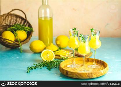 Limoncello with thyme in three grappas wineglass in wooden tray, fresh lemon in basket on light concrete table. Artistic still life on light background.