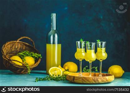 Limoncello with thyme in three grappas wineglass in wooden tray, fresh lemon in basket. Artistic still life on dark blue background