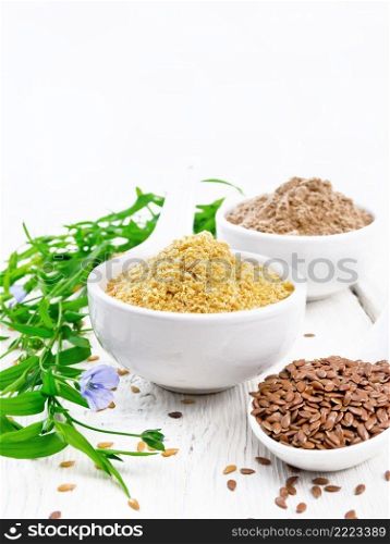 Linen bran and flour in two white bowls, seeds in spoons, leaves and blue flowers of flax on the background of a wooden board