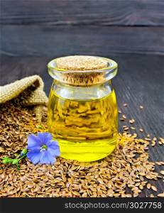 Linen oil in a glass jar with seeds in a bag and on a table, blue flax flower on a background of a dark wooden board. Oil linenseed with flower and seeds on dark board