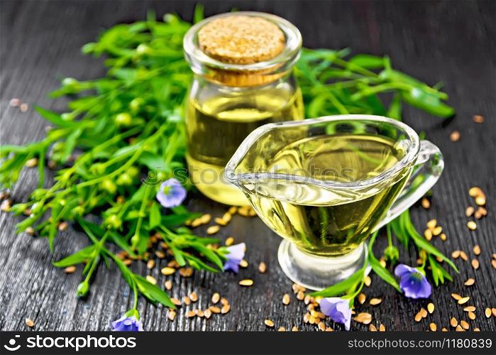 Linseed oil in a glass jar and gravy boat with seeds, leaves and flax flowers on a wooden board background