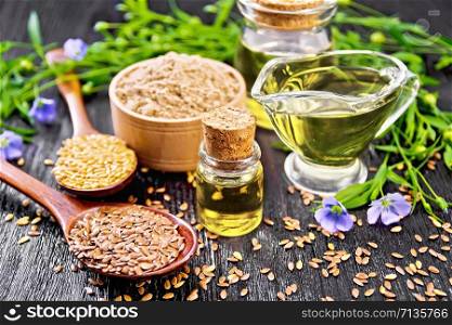 Linseed oil in two glass jars and a gravy boat with white and brown flax seeds in spoons, flour in a bowl, leaves and flowers on black wooden board background