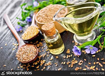 Linseed oil in two glass jars and a sauce boat with white and brown flax seeds in spoons, flour in a bowl, leaves and blue flowers on wooden board background