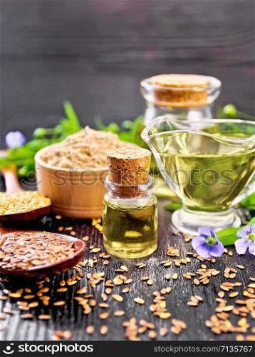 Linseed oil in two glass jars and a sauce boat with white and brown flax seeds in spoons, flour in a bowl, leaves and flowers on wooden board background