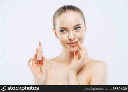 Lip care concept. Pensive beautiful woman with dark hair, poses with cosmetic product, has minimal makeup, looks sensually aside, poses topless, has well cared body, isolated on white background
