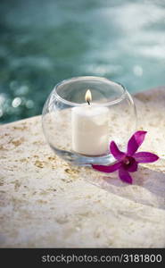 Lit candle in glass bowl with purple orchid next to pool.