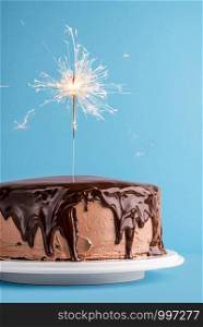 Lit sparkler on birthday cake with cocoa buttercream and melted chocolate glaze, on blue background. New year concept with festive cake and firework.
