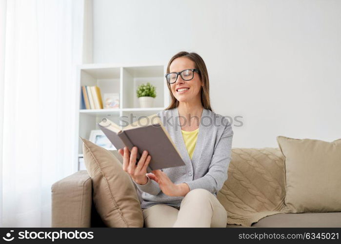 literature, education and people concept - happy middle-aged woman in glasses reading book at home. happy woman in glasses reading book at home