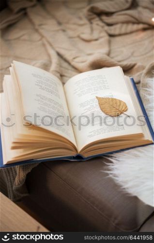 literature, reading and poetry concept - book with autumn leaf on page on sofa at home. book with autumn leaf on page on sofa at home