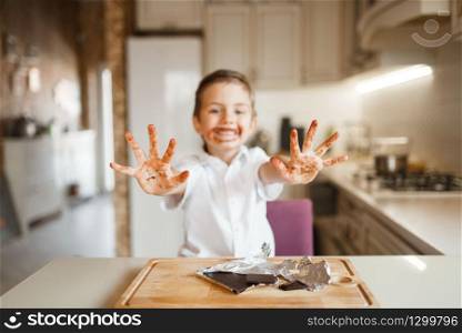 Litte boy shows hands stained with melted chocolate. Kid cooking on the kitchen. Happy child prepares sweet dessert at the counter
