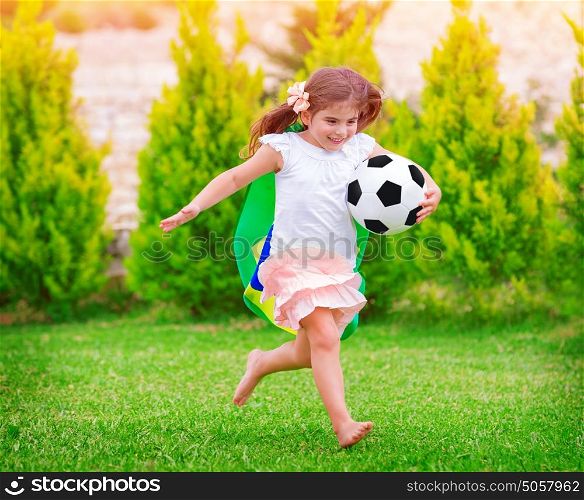 Little active football fan running on the green grass field with ball, young supporter of Brazilian football team with national flag
