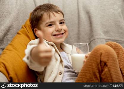 Little adorable boy sitting on the couch at home and drinking milk. Fresh milk in glass, dairy healthy drink. Healthcare, source of calcium, lactose. Preschool child with casual clothing. Thumbs up. Little adorable boy sitting on the couch at home and drinking milk. Fresh milk in glass, dairy healthy drink. Healthcare, source of calcium, lactose. Preschool child with casual clothing. Thumbs up.