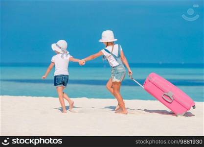 Little adorable girls with big luggage in hands walking on tropical beach to spend summer vacation. Little tourists girls with big suitcase on tropical white beach