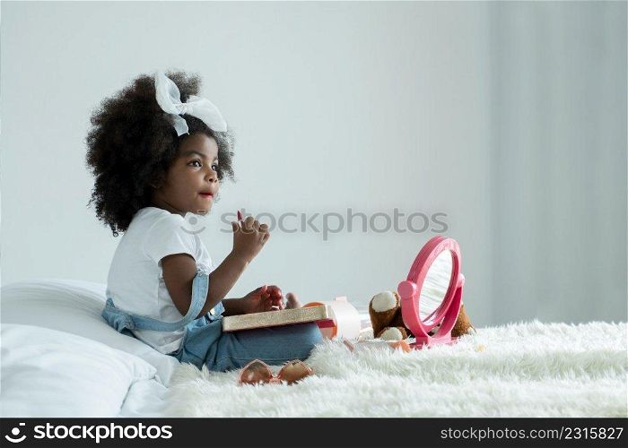 Little African girl playing makeup with mother"s cosmetics. Adorable kid painting her lips with red lipstick and looking in the mirror with joy in bedroom at home