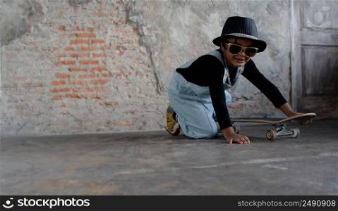 Little African kid boy wear cool gold sunglasses, hat, necklace chain, jeans bib and sneaker smiling and have fun lying on skateboard on cement floor and brick wall on background