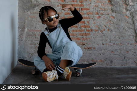 Little African kid boy wear cool gold sunglasses, necklace chain, jeans bib and sneaker smiling and sitting on skateboard with doing hip hop gesture on cement floor and brick wall on background